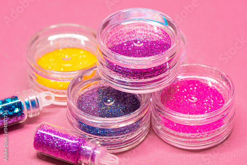 Collection of small plastic containers filled with various colors of glitter, on a pink surface. The vibrant and diverse colors of the glitter. Crafts, makeup, decoration concepts photo