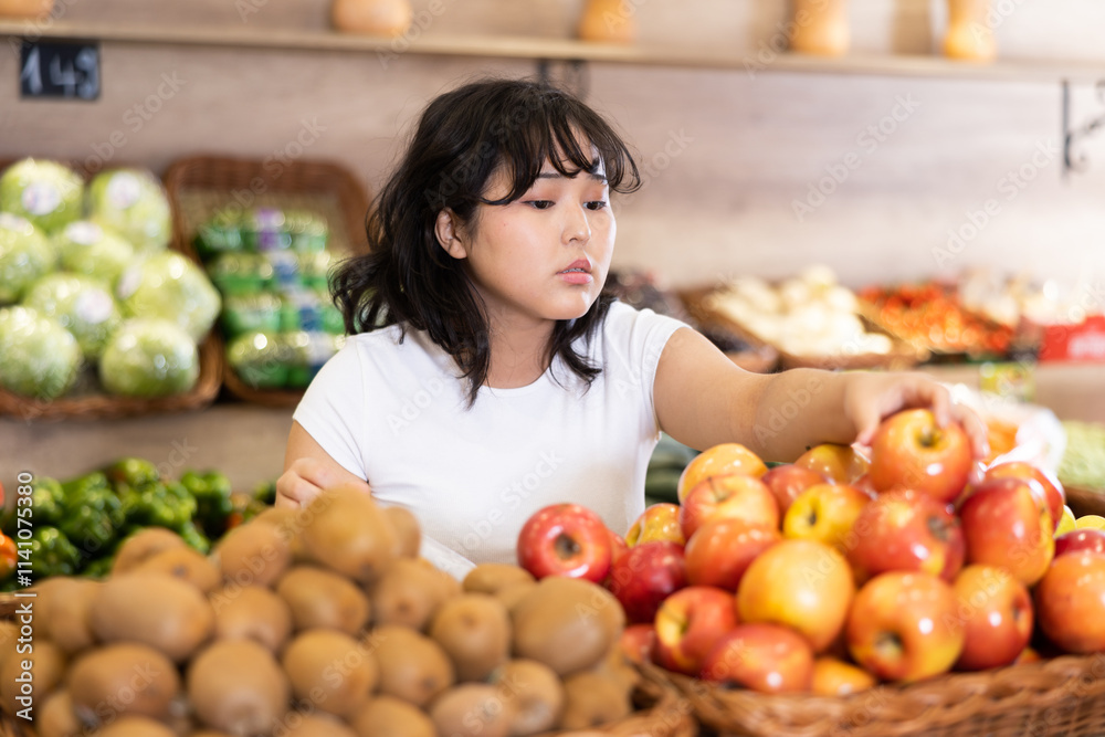 Interested young asian girl shopping for organic fruits in farm store, choosing ripe apples from wicker box on produce display.. Interested young asian girl shopping for organic fruits in farm store, choosing ripe apples from wicker box on produce display..