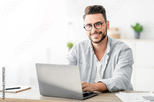 Portrait of freelancer man sitting at desk with laptop computer at home office, looking and smiling at camera, free space. Happy entrepreneur guy enjoying remote work and self-employment