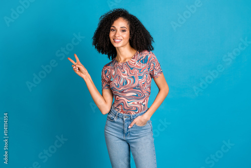 Stylish young woman with curly hair wearing a trendy t-shirt makes a peace gesture against a vivid blue backdrop