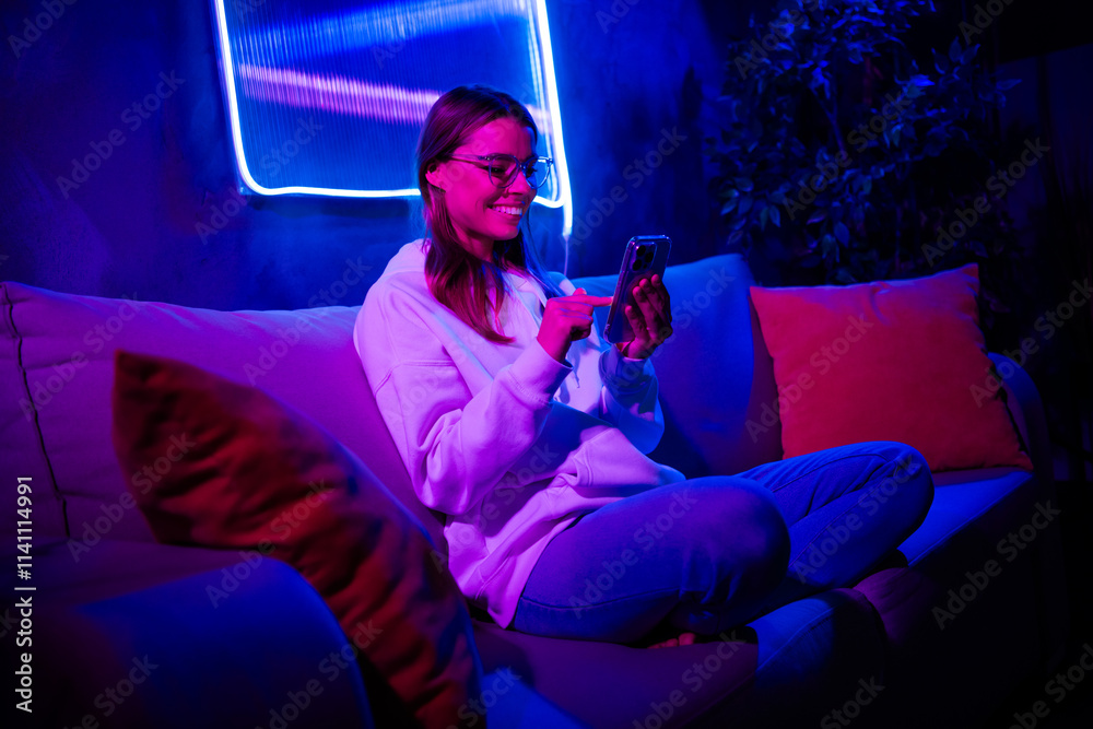 Naklejka premium Full length photo of gamer lovely blonde young lady hold gadget sit sofa blogging neon light neon purple late night room indoors