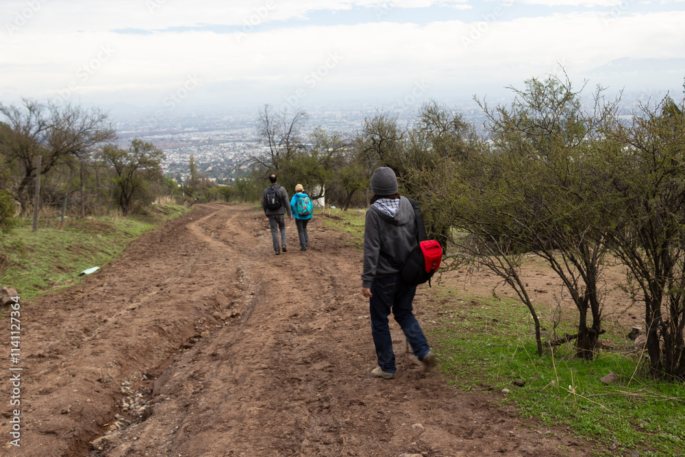 Dos personas con ropa de otoño por un sendero de tierra, atrás queda una persona con gorro gris mirándolos alejarse, al fondo la ciudad de Santiago, Chile Dos personas con ropa de otoño por un sendero de tierra, atrás queda una persona con gorro gris mirándolos alejarse, al fondo la ciudad de Santiago, Chile