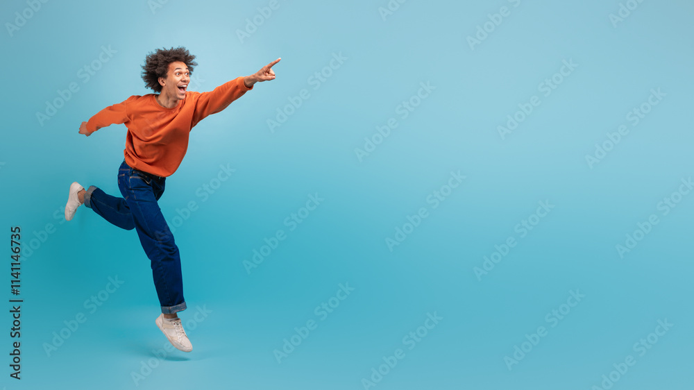 A young African American man with curly hair is energetically running and pointing at copy space, dressed in a casual orange sweatshirt and jeans. A young African American man with curly hair is energetically running and pointing at copy space, dressed in a casual orange sweatshirt and jeans.