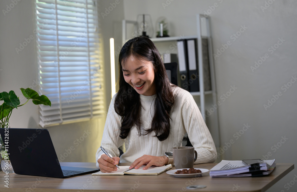 Young beautiful businesswoman smiling and writing notes on notebook while working on laptop computer at her home office desk at night, work from home, telecommuting concept Young beautiful businesswoman smiling and writing notes on notebook while working on laptop computer at her home office desk at night, work from home, telecommuting concept