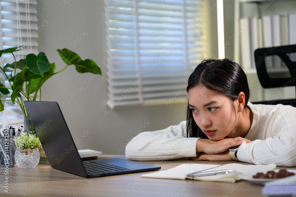 Young asian freelancer is staring at her laptop screen with a bored expression, resting her chin on her hands, showing signs of tiredness and lack of motivation while working from home Young asian freelancer is staring at her laptop screen with a bored expression, resting her chin on her hands, showing signs of tiredness and lack of motivation while working from home