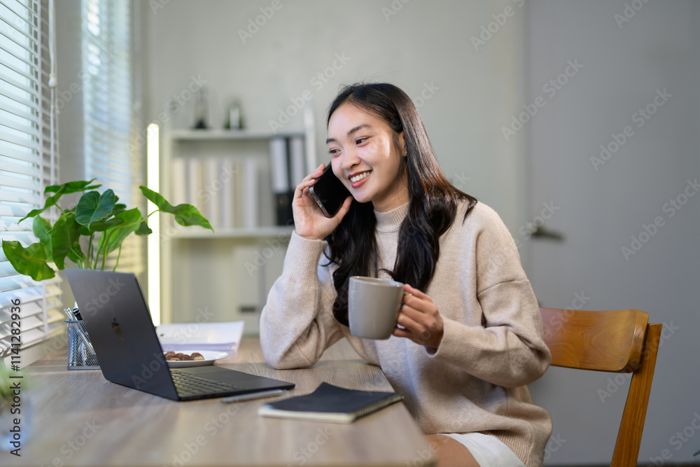 Young professional woman is working remotely from home, enjoying a cup of coffee while having a phone conversation and using laptop, showcasing modern work-life balance Young professional woman is working remotely from home, enjoying a cup of coffee while having a phone conversation and using laptop, showcasing modern work-life balance