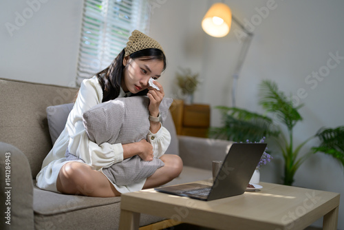 Young asian woman sitting on the sofa, hugging a pillow tightly while wiping away tears, watching a sad movie or receiving distressing news on her laptop