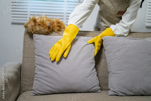 Housekeeper wearing yellow gloves is arranging pillows on a sofa in the living room, ensuring a tidy and comfortable environment for residents or guests
