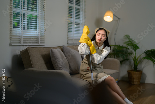 Young asian housekeeper wearing yellow gloves and an apron, resting on the sofa with a mop after a long day of cleaning, feeling tired and completely exhausted from her chores