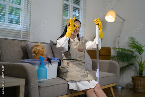 Smiling housekeeper wearing yellow gloves and apron gesturing ok sign while holding cleaning mop sitting on sofa in living room with cleaning supplies and cute dog