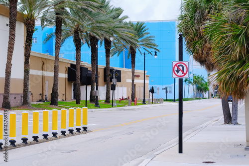 Street with palm trees, buildings, yellow traffic barriers, a no U-turn sign, and a sidewalk lined with trimmed greenery. photo