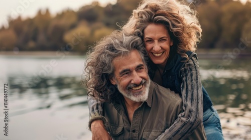 Happy middle-aged couple hugging each other and smiling at the camera, with sunlight shining on their faces, a lake in the background and a grassy meadow.