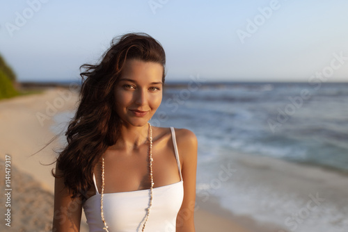 A smiling woman in a white dress enjoys a peaceful moment on the beach during sunset, with the ocean behind her. Perfect for summer, travel, beach lifestyle, and relaxation promotions.