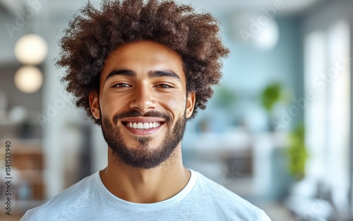 A man with a beard is smiling and wearing a t-shirt in a blurred room background