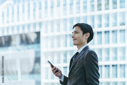 高層ビルのあるオフィス街で自身を持った表情でスマートフォンを持ち遠くを眺める30代の男性 photo