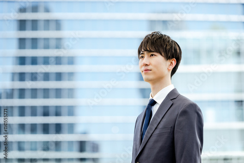 オフィスビルのガラスの建物の前で真面目な表情で遠くを見つめる20代のスーツを着た新入社員の男性