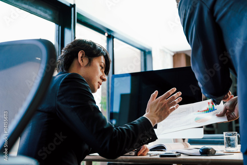 オフィスのデスクでパソコンモニターを前に部下から見せられた資料に指示を与える管理職の上司の男性