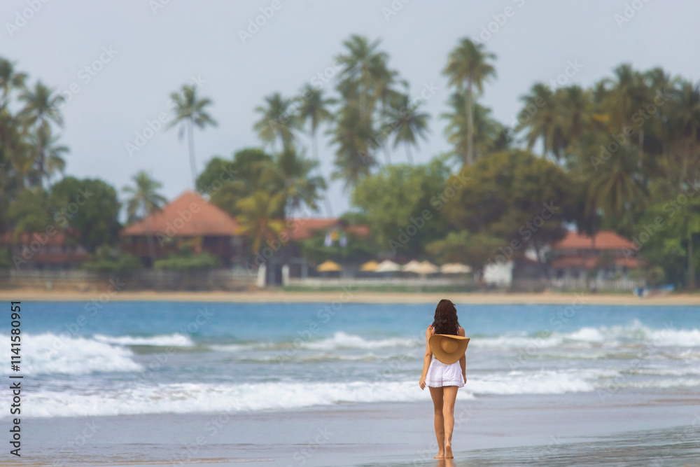 Obraz premium A woman walks barefoot along a tropical beach in Dickwella, Sri Lanka, holding a sun hat as palm trees and ocean waves surround her. Perfect for travel, vacation, and tropical lifestyle concepts or
