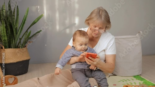 A grandmother and her young grandson sit comfortably on a floor mat. They share a joyful interaction as the grandmother helps the child explore the smartphone, promoting bonding and learning