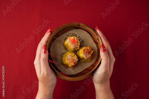 Asian women hand serving shumai dim sum using woven bamboo steamer plate. Steamed dumplings for Chinese New Year party, isolated on red background.