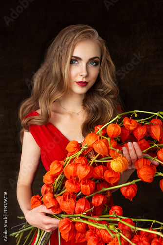 Close-up of beautiful young blonde girl with long light red hair in long red dress holding big bouquet of dried orange physalis flowers on black background. Studio photography. Autumn art portrait. photo