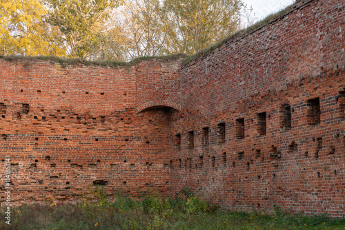 Brick ruins of a medieval castle wall with arched elements and rectangular openings, surrounded by autumn foliage. A historic structure showcasing weathered red brickwork and aged architecture. photo