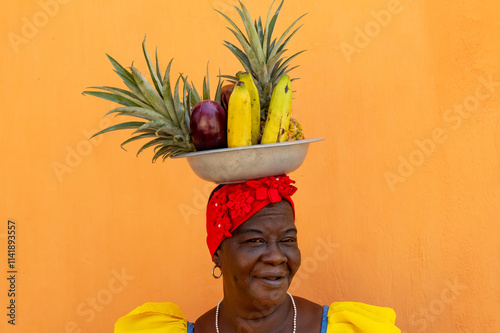 Woman In A Colorful Colombian Dress Balancing A Bowl Of Tropical Fruits Against An Orange Wall photo