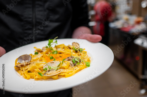 A chef holds a plate of fresh fettuccine pasta garnished with herbs and clams, showcasing culinary artistry in a stylish kitchen setting. photo