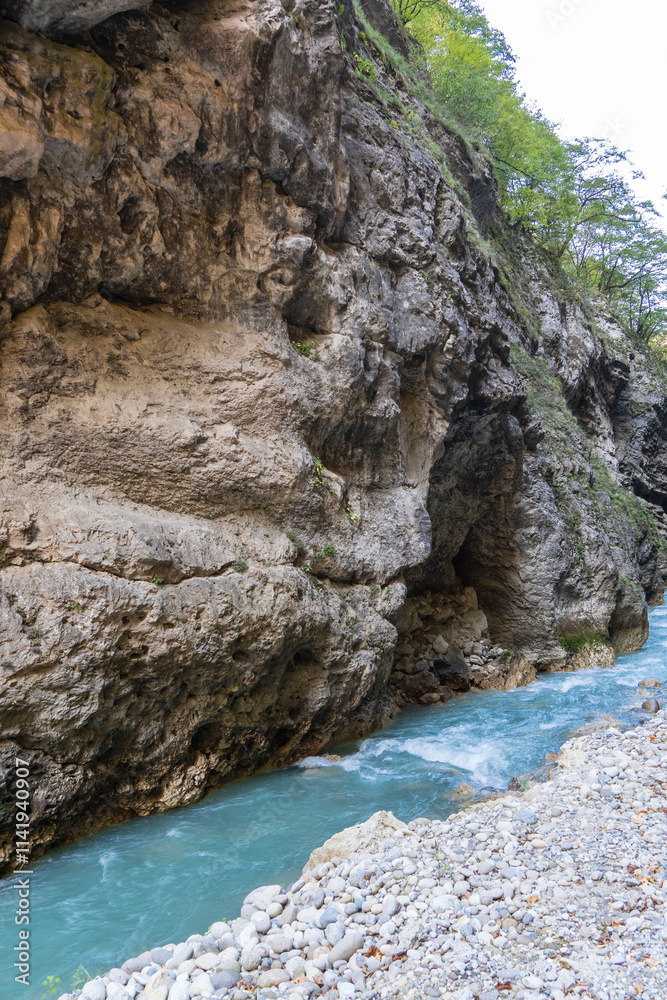 Steep cliffs hang over mountain river with emerald-clear water. Stormy Chegem River flows in deep crevice along bottom of Chegem Gorge. In depths of gorge, semi-darkness reigns. Steep cliffs hang over mountain river with emerald-clear water. Stormy Chegem River flows in deep crevice along bottom of Chegem Gorge. In depths of gorge, semi-darkness reigns.