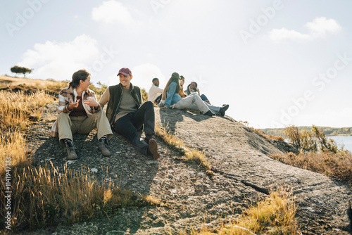 Happy young man talking with female friend while sitting on hill at sunny day photo