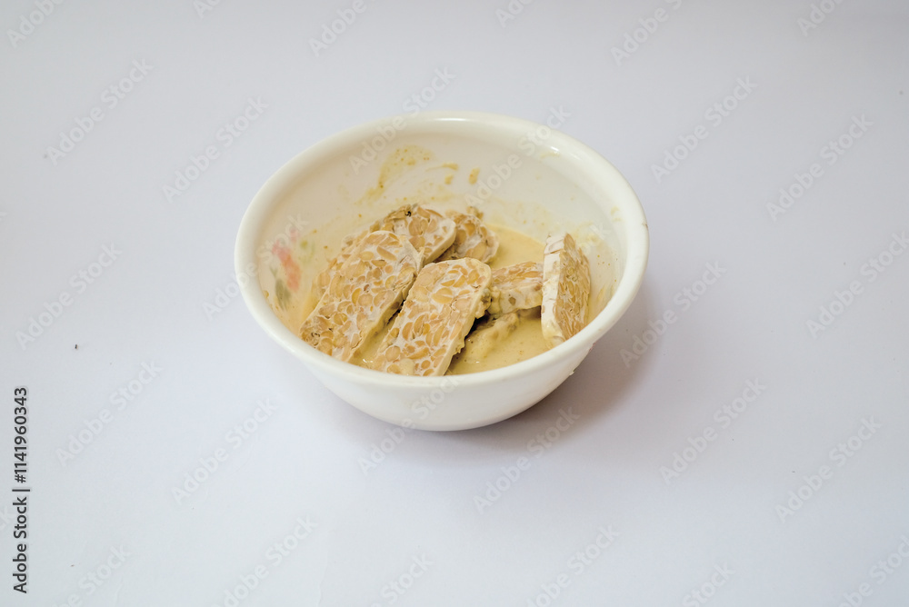 Raw tempeh that has been cut in a white bowl containing dough, with a white background. Raw tempeh that has been cut in a white bowl containing dough, with a white background.