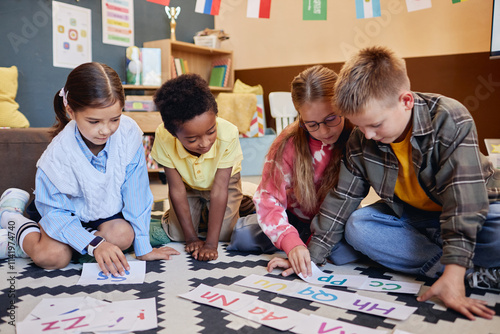 Group of young schoolkids playing alphabet game on floor and laying cards while learning English together photo