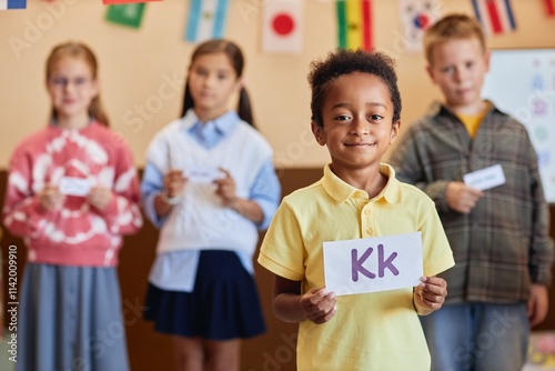 Waist up portrait of African American boy holding letter card and looking at camera with group of children learning English in background photo