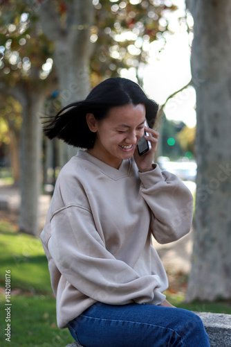 Smiling Asian woman talking on the phone while sitting on a bench in a sunny park surrounded by trees, vertical image photo