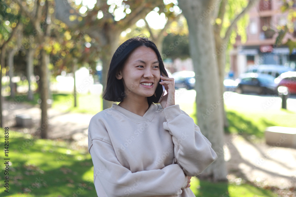Young Asian woman smiling while having a phone call in a sunny urban park with trees and cars in the background Young Asian woman smiling while having a phone call in a sunny urban park with trees and cars in the background