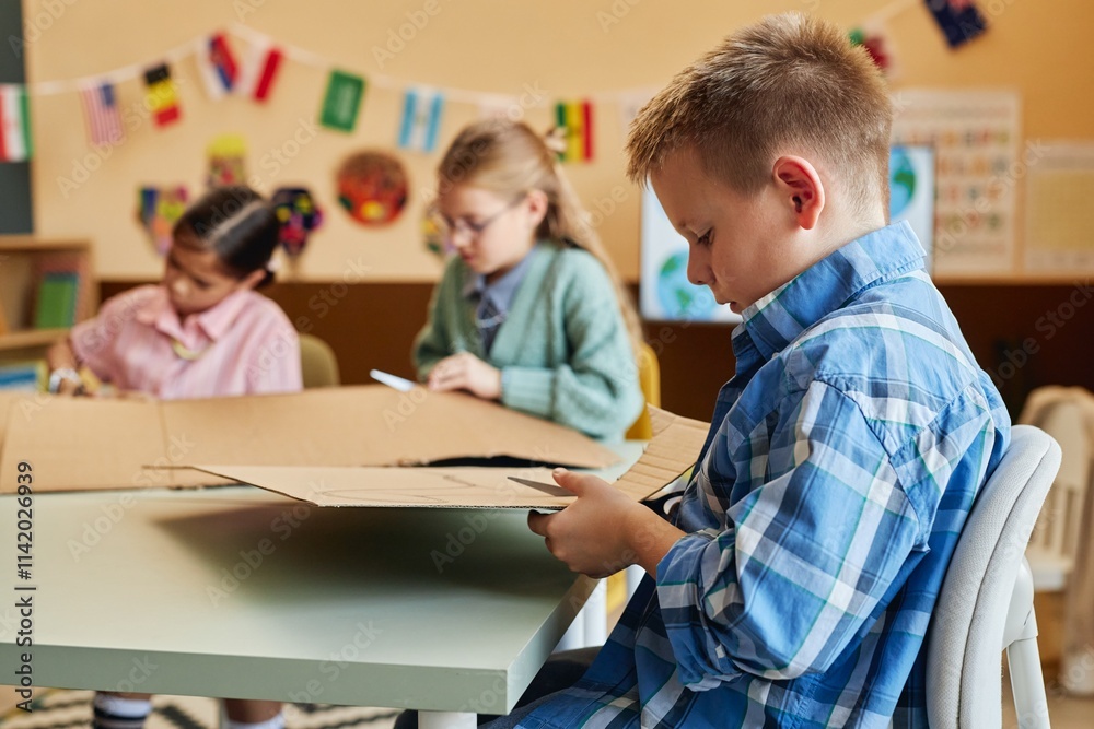 Side view portrait of blonde young boy cutting cardboard while enjoying art and craft class for children copy space Side view portrait of blonde young boy cutting cardboard while enjoying art and craft class for children copy space