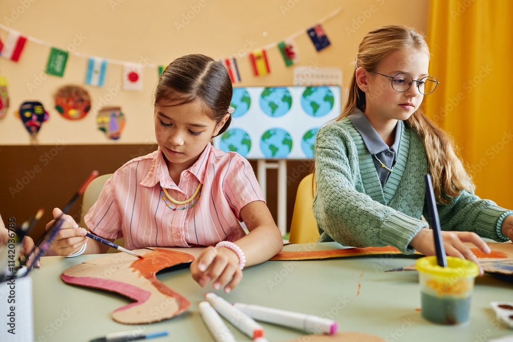 Portrait of two young girls painting dinosaur figures made of cardboard while enjoying creative activity in colorful classroom copy space Portrait of two young girls painting dinosaur figures made of cardboard while enjoying creative activity in colorful classroom copy space