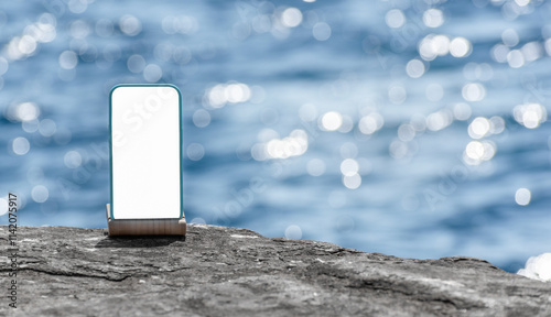 Mobile phone with white blank screen mock-up on rocky seashore, blurred blue water background and copy space. Phone device mockup for text, design, advertising, travel insurance, resort real estate