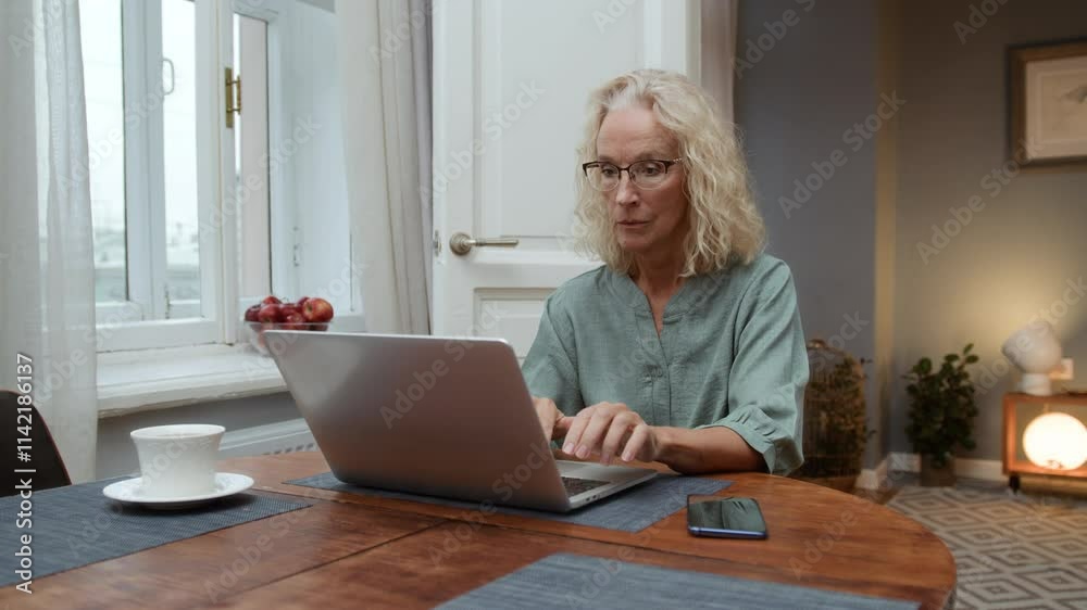 Senior elegant woman sits at table typing on keyboard. Cheerful aged woman working remotely using laptop. Older focused businesswoman browsing online. Female elegant writer enjoying creative process Senior elegant woman sits at table typing on keyboard. Cheerful aged woman working remotely using laptop. Older focused businesswoman browsing online. Female elegant writer enjoying creative process
