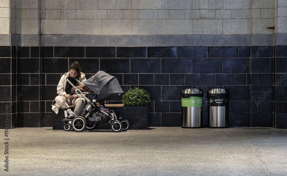 Portrait of Woman using her smartphone while sitting on the bench next to beby in stroller(baby carriage), plant pot and Stainless steel trash cans and recycle bins at resting area inside the shopping
