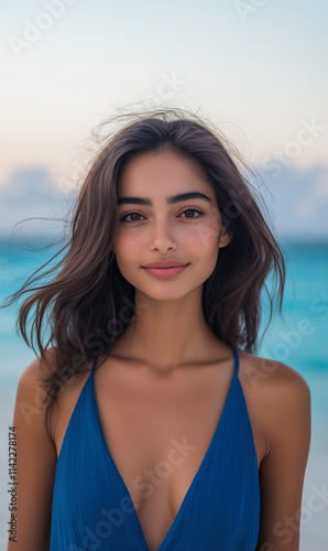 An extremely beautiful and attractive woman with medium-length hair, wearing an electric blue sleeveless dress, standing on the beach in front of turquoise water in the Maldives