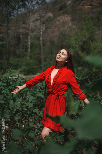the girl stands against a background of green foliage and trees, with her eyes closed and her hands raised, touching her head. Her facial expression is calm and relaxed. photo