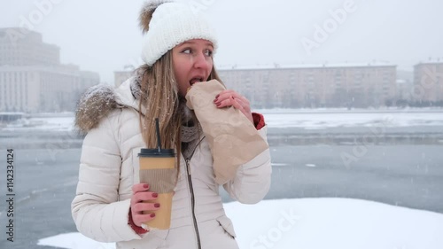  A young woman eats a sweet pretzel outside during a snowfall. Street food. Winter.