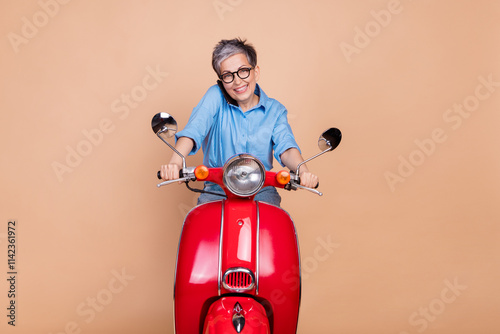 Trendy senior woman enjoying leisure riding red scooter against beige background photo