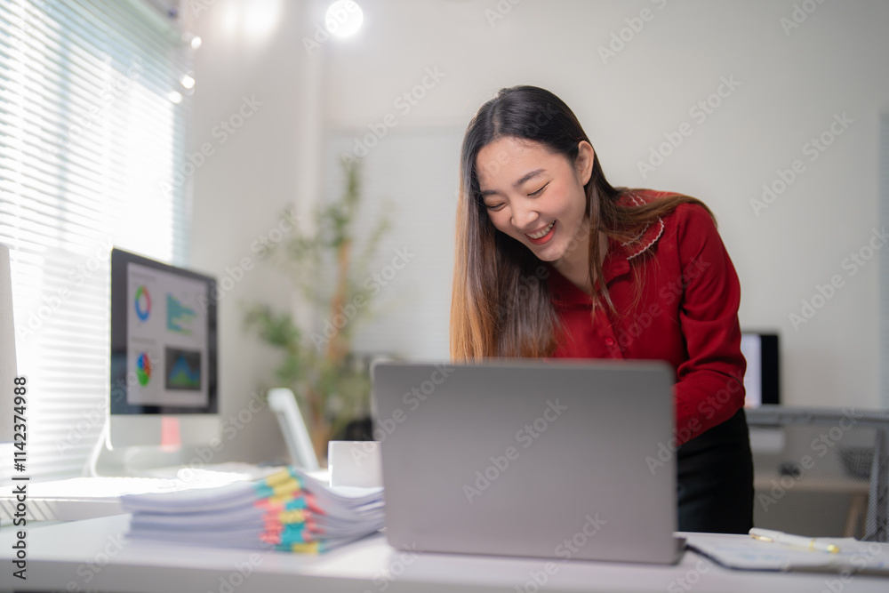 Naklejka premium Asian businesswoman is standing at her desk, laughing while working on her laptop, with a pile of paperwork and a desktop computer in the background