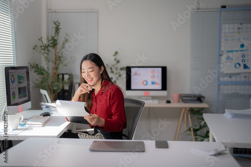 Young asian businesswoman holding pen and reading financial documents, smiling and working at office desk with computer showing graphs, startup business concept