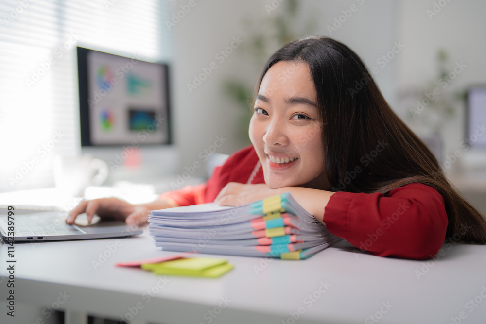 Asian businesswoman is smiling while resting on a stack of documents with colored sticky notes at her modern office desk, working on a laptop computer with a desktop screen in the background Asian businesswoman is smiling while resting on a stack of documents with colored sticky notes at her modern office desk, working on a laptop computer with a desktop screen in the background