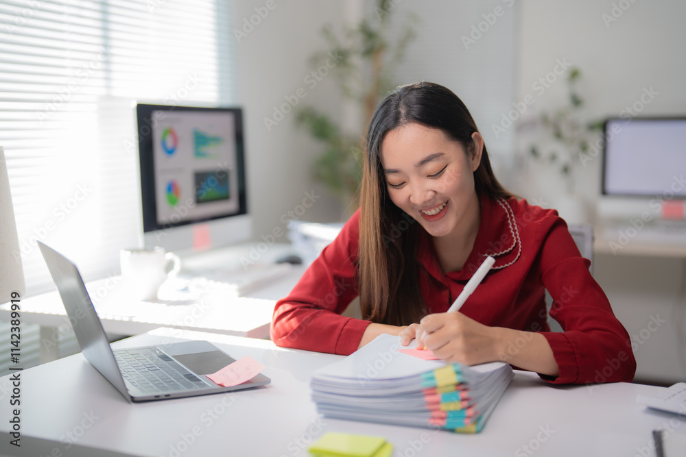 Young asian businesswoman smiling and working at her desk in a modern office, using a laptop and taking notes on documents, enjoying her productive workday Young asian businesswoman smiling and working at her desk in a modern office, using a laptop and taking notes on documents, enjoying her productive workday