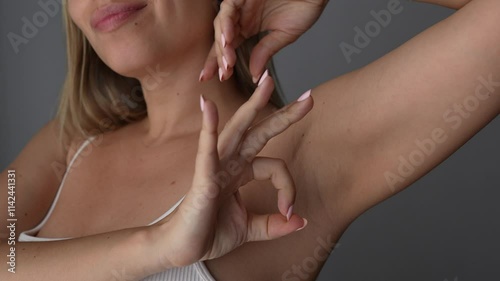 Young blonde woman showing okay gesture against the armpit on dark grey background. Deodorant for getting rid, masking and blocking the smell of sweat. The result of laser hair removal. OK sign