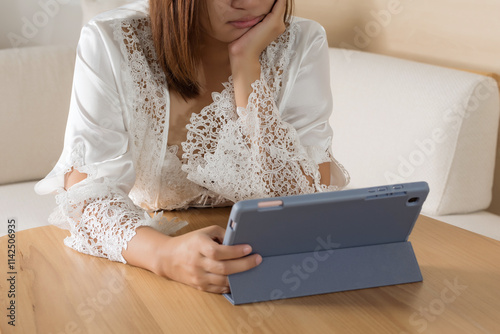A woman in white silk sleepwear reads and checks her work on a tablet at night.
